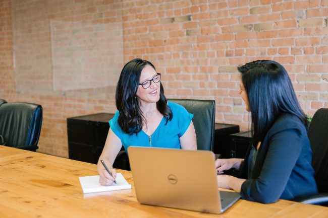 two women sitting at a desk, talking. meeting